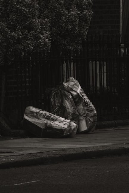 A narrow, urban alleyway cluttered with overflowing rubbish and discarded materials. In the foreground, a large black garbage bag is prominently positioned on the pavement, appearing to be weathered a